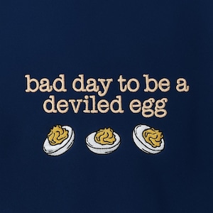 May include: A navy blue design featuring the embroidered text "bad day to be a deviled egg." Below the text are three embroidered deviled eggs, each with a white egg white and yellow filling.