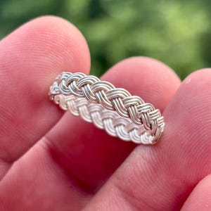 May include: A silver-toned ring featuring a continuous braided or woven metal pattern that encircles the entire band. The surface of the ring displays a polished metallic finish with intricate, repeating diagonal strands that create a textured look.