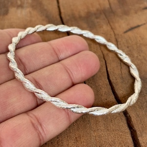 Può includere: Un braccialetto rigido color argento con un design a corda intrecciata. Il braccialetto è realizzato in metallo lucido e ha una superficie strutturata. Il gioiello è esposto su una superficie di legno.