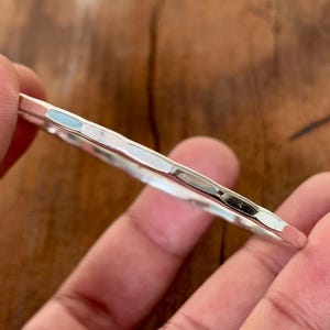 Puede incluir: Una pulsera de plata martillada. La pulsera tiene una superficie texturizada con pequeñas facetas irregulares que captan la luz. La pulsera se sostiene entre dos dedos, destacando su diseño simple y elegante. El diámetro es de aproximadamente 6,5 cm.
