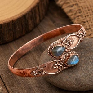Può includere: Un bracciale a fascia in rame dal design aperto con due pietre preziose di labradorite a forma di goccia. La fascia metallica presenta una finitura martellata e testurizzata ed è decorata con piccole sfere e motivi a spirale alle estremità.
