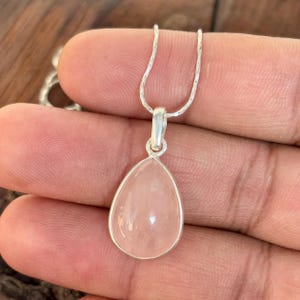 May include: A silver pendant necklace with a teardrop-shaped rose quartz gemstone. The pale pink stone is set in a silver bezel and hangs from a delicate silver chain. The necklace is displayed against a natural wood background.