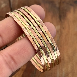 Pode incluir: Um conjunto de pulseiras de metal martelado em tons de cobre e dourado. As pulseiras têm uma superfície texturizada e são empilhadas. Os tons quentes do metal criam um visual rústico e elegante. As pulseiras são um tipo de joia.