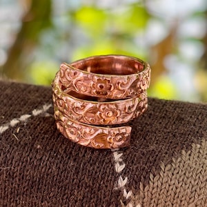 Può includere: Un anello color rame con un motivo floreale si avvolge attorno al dito. L'anello ha una superficie strutturata con incisioni dettagliate di fiori e foglie. L'anello è esposto su una superficie a maglia marrone.
