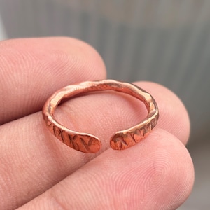 Può includere: Un anello in rame con design a fascia aperta, caratterizzato da una superficie testurizzata e martellata con piccole rientranze ripetute lungo l'intera circonferenza esterna. Il metallo ha una calda finitura metallica bruno-rossastra e una forma arrotondata.