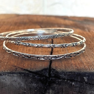 May include: A set of three silver-toned metal bangles featuring intricate embossed floral and leaf patterns. Each bangle has a polished finish with darkened recessed areas that highlight the detailed botanical designs along the entire circumference.