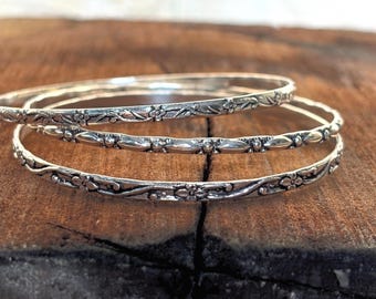 May include: A set of three silver-toned metal bangles featuring intricate embossed floral and leaf patterns. Each bangle has a polished finish with darkened recessed areas that highlight the detailed botanical designs along the entire circumference.