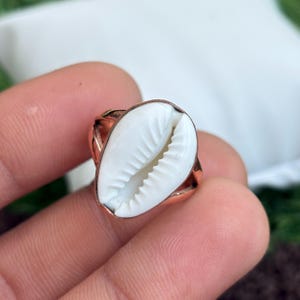 Puede incluir: Un anillo de color cobre con una concha blanca. La concha tiene forma ovalada con una superficie texturizada y está engastada en una banda de color cobre. El anillo es una joya.