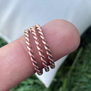 Könnte beinhalten: Drei kupferfarbene, gedrehte Seilringe. Die Ringe sind auf einem Finger gestapelt und zeigen ihr detailliertes, strukturiertes Design. Die Ringe haben einen warmen, metallischen Glanz, der das Licht reflektiert. Der Hintergrund ist verschwommen, mit Andeutungen von Grün und Weiß.