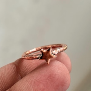 Può includere: Un anello color rame caratterizzato da una fascia martellata e testurizzata, con un ciondolo a forma di stella a cinque punte liscia centrato sul davanti. Il metallo ha una finitura calda con sottili variazioni di luce sulla superficie.