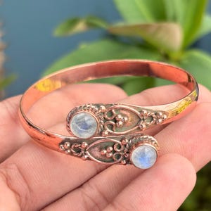 Può includere: Un bracciale a fascia in rame dal design aperto, caratterizzato da due cabochon rotondi in pietra di luna iridescente. Le estremità in metallo sono decorate con complessi motivi a volute in rilievo e piccole perline, creando una finitura metallica.