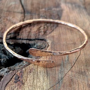 Può includere: Un bracciale a polsino in rame martellato con un design aperto. Il bracciale ha una superficie strutturata e estremità arrotondate. Il caldo colore ramato contrasta con lo sfondo in legno. Questo gioiello artigianale è progettato per essere indossato al polso.