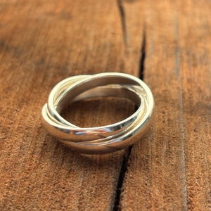 Peut inclure: Une bague argentée composée de trois anneaux entrelacés, formant une boucle continue. La bague, en métal brillant, repose sur une surface en bois brun texturé. Le design de la bague est simple et élégant.