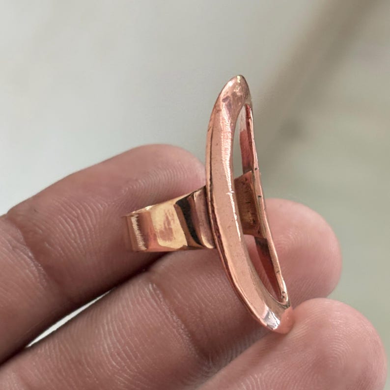 Pode incluir: Um anel de cor cobre com um design oval aberto, alto e vertical. A superf&iacute;cie met&aacute;lica tem um acabamento polido e reflexivo com uma banda curva e lisa que se conecta &agrave; pe&ccedil;a central alongada e oca, criando um visual met&aacute;lico marcante.