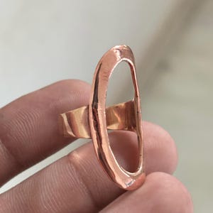 Pode incluir: Um anel de tom cobre com uma estrutura grande em forma de oval alongada. A superf&iacute;cie met&aacute;lica tem um acabamento liso e polido com uma qualidade reflexiva. A banda est&aacute; ligada ao lado da estrutura oval vertical, criando um design aberto e arrojado.
