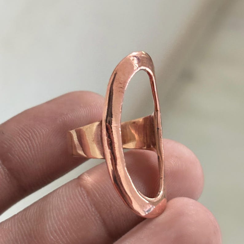 Pode incluir: Um anel de tom cobre com uma estrutura grande em forma de oval alongada. A superf&iacute;cie met&aacute;lica tem um acabamento liso e polido com uma qualidade reflexiva. A banda est&aacute; ligada ao lado da estrutura oval vertical, criando um design aberto e arrojado.