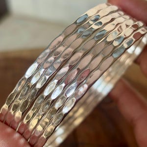 Pode incluir: Uma pilha de pulseiras de prata com textura martelada. As pulseiras são feitas de metal brilhante e refletivo e têm uma superfície ligeiramente irregular. As pulseiras são empilhadas, criando um efeito em camadas. Joias para uso diário.