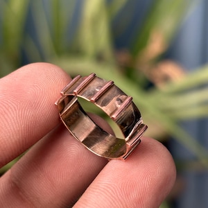 Könnte beinhalten: Ein kupferfarbener Metallring mit glattem Innenband und einer Außenfläche, die durch eine Reihe gleichmäßig verteilter, vertikaler erhabener Rippen gekennzeichnet ist, die sich um den gesamten Umfang des kreisförmigen Schmuckstücks erstrecken.