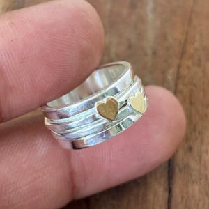 Puede incluir: Un anillo de plata con dos corazones dorados. El anillo tiene un diseño en capas con un acabado pulido. El anillo es una joya.