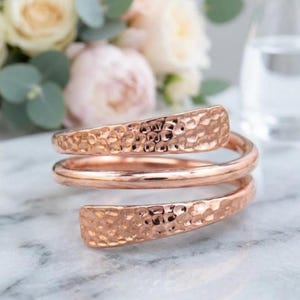 Op de afbeelding: Een roségoudkleurige bypass-ring met een spiraalontwerp, twee platte uiteinden met een gehamerde textuur en een gladde, afgeronde centrale band. Het metaal heeft een gepolijste, reflecterende afwerking die het licht opvangt, wat zorgt voor een duidelijk contrast tussen de getextureerde uiteinden en de strakke, doorlopende cirkelvormige structuur van de ring.