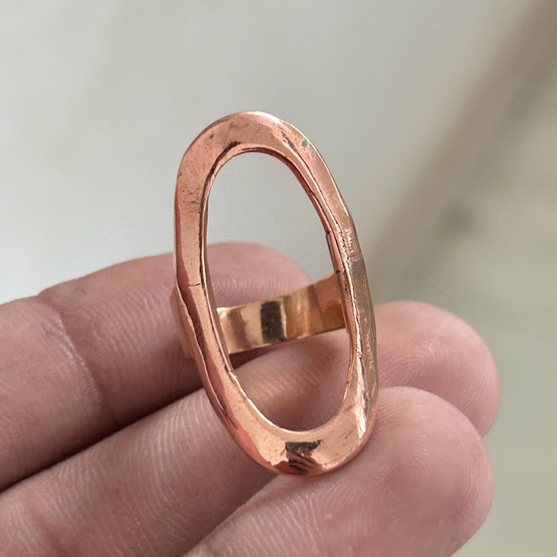 Pode incluir: Um anel de cobre com um topo grande de formato oval alongado com um acabamento martelado e texturizado. A banda &eacute; lisa e conecta-se &agrave; base da estrutura oval vertical, criando um design aberto e arrojado com um tom castanho-avermelhado.