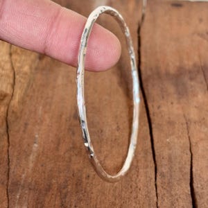 Pode incluir: Uma pulseira de prata martelada. A pulseira tem uma superfície texturizada e uma forma circular. A prata é brilhante e reflexiva, e a pulseira é mostrada contra uma superfície de madeira castanha.