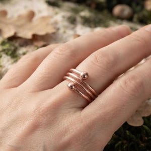 May include: A copper-colored ring featuring a multi-band design with three parallel wires that wrap around the finger. The ends of the outer wires are finished with small, smooth spherical beads, creating a layered and continuous metallic appearance.