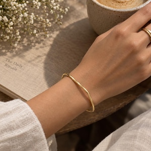 Pode incluir: Uma pulseira fina de metal em tom dourado com uma silhueta ondulada orgânica e subtil. A pulseira apresenta um acabamento liso e polido e um design contínuo e flexível que repousa confortavelmente à volta do pulso, exibindo uma estética simples.