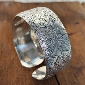 Könnte beinhalten: Ein breites, silbernes Armreif-Armband mit einem komplizierten, geprägten Blumenmuster. Das offene Design ermöglicht ein einfaches Tragen. Das Armband wird auf einer Holzoberfläche präsentiert, die seinen metallischen Glanz hervorhebt.