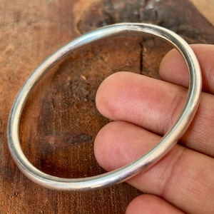 Può includere: Un braccialetto ovale color argento. Il braccialetto liscio sembra essere in metallo. È tenuto dalle dita di una persona, con una superficie di legno sullo sfondo.