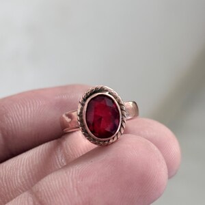 Könnte beinhalten: Ein Ring mit einem zentralen, ovalen und facettierten tiefroten Edelstein. Der Stein ist in eine gedrehte, seilartige Metallfassung eingefasst. Die Ringschiene ist glatt und metallisch, mit einem kleinen, klaren Akzentstein an der Seite.