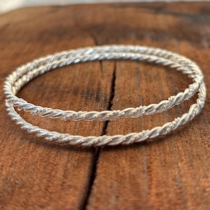 Puede incluir: Dos pulseras plateadas con un diseño de cuerda trenzada. Las pulseras se exhiben sobre una superficie de madera marrón. Las pulseras tienen un aspecto brillante y metálico y una superficie texturizada. El diseño es un patrón continuo en espiral.