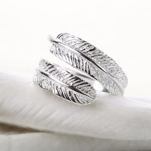 May include: A silver ring with a feather design. The ring features two detailed feather shapes that wrap around the finger. The ring is displayed on a white surface, highlighting the intricate details of the feather design.