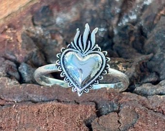 May include: A silver-toned ring featuring a heart-shaped design with a flame-like element at the top. The heart is surrounded by a scalloped border. The ring is set against a textured, brown background.