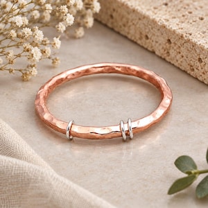 May include: A circular bangle bracelet with a hammered copper finish. The bracelet features three small, smooth silver-toned rings positioned along the bottom section of the band, creating a contrast between the copper and silver metal tones.