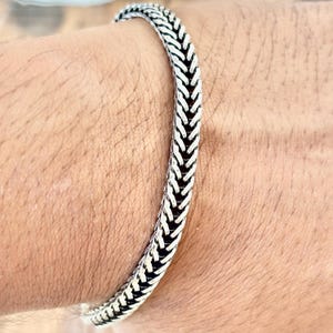May include: A silver bracelet with a woven pattern. The bracelet is made of a shiny metal and has a textured surface. The bracelet is worn on a wrist.