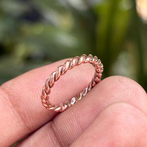 Könnte beinhalten: Ein dünner, kreisförmiger Ring mit einer durchgehenden gedrehten Seilstruktur. Das Metall hat einen warmen, rötlich-kupferfarbenen Ton mit einer polierten Oberfläche, die das Licht entlang des Spiralmusters des Bandes reflektiert.