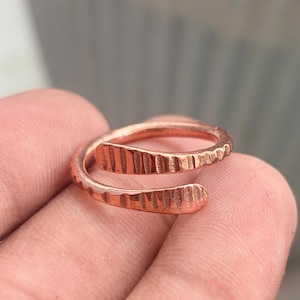 Op de afbeelding: Een koperen ring met een open bypass-ontwerp en een getextureerde, gehamerde afwerking. De band heeft twee overlappende uiteinden die elkaar niet raken, wat zorgt voor een spiraalvormig uiterlijk met zichtbare inkepingen op de buitenkant.