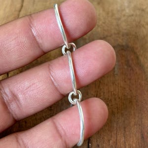 May include: A close-up of a silver chain with three elongated, curved links connected by small, circular clasps. The chain is held against a blurred background, highlighting the polished metal.