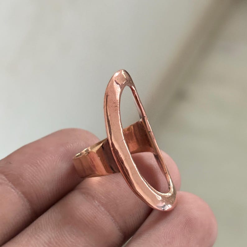Pode incluir: Um anel de cobre com uma pe&ccedil;a central grande, alongada e de formato oval vertical, com uma superf&iacute;cie lisa e polida. A banda est&aacute; ligada ao lado da estrutura oval, criando um design aberto com um tom met&aacute;lico avermelhado e quente.