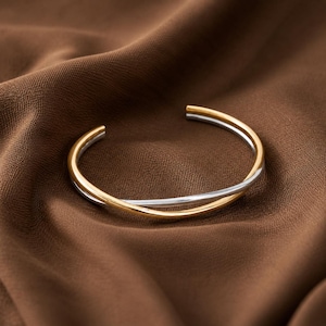 Peut inclure: Un bracelet manchette bicolore composé d'un anneau doré poli entrelacé avec un anneau argenté lisse. La conception circulaire ouverte crée un effet superposé avec des finitions métalliques contrastées qui captent la lumière.