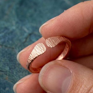 Peut inclure: Une bague ouverte de couleur cuivre avec une finition de surface texturée et martelée. L'anneau est circulaire avec deux extrémités arrondies et légèrement évasées qui ne se rejoignent pas, créant un espace. Le métal a un ton brun-rouge chaud.