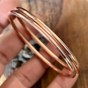 以下が含まれることがあります： 3つの銅色のバングルが重ねられています。滑らかで磨かれた金属は光を反射し、ハイライトと影を作り出しています。バングルは丸く、しっかりとした素材でできているようです。全体的な美しさはシンプルでエレガントです。