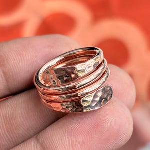 Op de afbeelding: Een koperen ring met een meerlaags wikkelontwerp en een gehamerde textuur. Het metaal heeft een gepolijste, reflecterende afwerking die de inkepingen op het oppervlak van de band benadrukt, wat een duidelijk visueel patroon van licht en schaduw creëert.