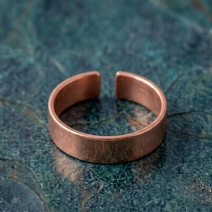 Könnte beinhalten: Ein schlichter Kupferring mit glatter, polierter Oberfläche und einfachem Banddesign. Der Ring weist an der Oberseite eine Öffnung auf, die eine Anpassung der Größe ermöglicht. Das Metall hat einen warmen, rotbraunen Farbton auf der Oberfläche.