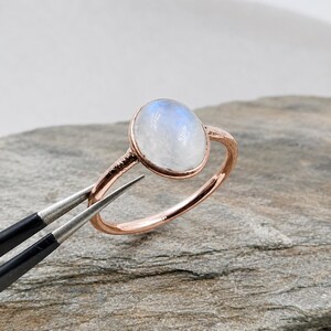 Könnte beinhalten: Ein Ring aus Roségold mit einem glatten, ovalen Mondstein-Cabochon in einer einfachen Metallfassung. Die Ringschiene hat eine polierte Oberfläche und ein schmales, abgerundetes Profil, das den irisierenden blau-weißen Glanz betont.