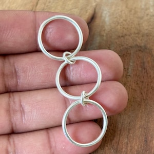 May include: A silver-colored pendant featuring three interconnected circular rings. The rings are linked together, creating a flowing, linear design. The pendant is made of a shiny, metallic material.