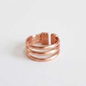 May include: A copper-colored metal ring featuring a triple-band design. The ring has an open-ended structure with a rectangular connector piece joining the three bands together at the back, creating a layered appearance with a smooth finish.