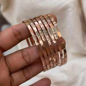 Może przedstawiać: Zestaw siedmiu bransoletek typu bangle z metalu w kolorze różowego złota o młotkowanej, teksturowanej powierzchni. Każda bransoletka ma cienki, zaokrąglony profil z odblaskowym wykończeniem, które chwyta światło, tworząc serię powtarzających się faset.
