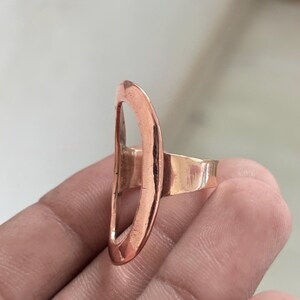 Pode incluir: Um anel de cobre com uma pe&ccedil;a central grande, vertical e em forma de oval aberta. O metal tem um acabamento met&aacute;lico quente de cor castanho-avermelhada com uma textura lisa e ligeiramente martelada. A banda &eacute; simples e est&aacute; ligada.